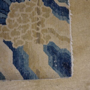 The edge of a flatweave rug with beige and blue abstract patterns rests on thick tan carpet.