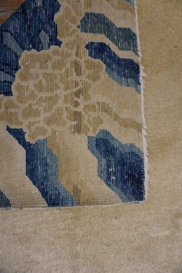 The edge of a flatweave rug with beige and blue abstract patterns rests on thick tan carpet.