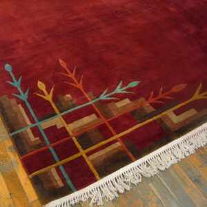 Deep red rug with a multicolored abstract border and white fringe rests on distressed wood flooring.