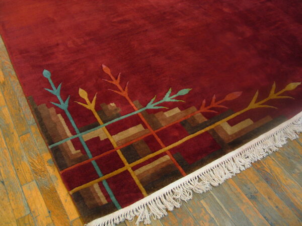 Deep red rug with a multicolored abstract border and white fringe rests on distressed wood flooring.