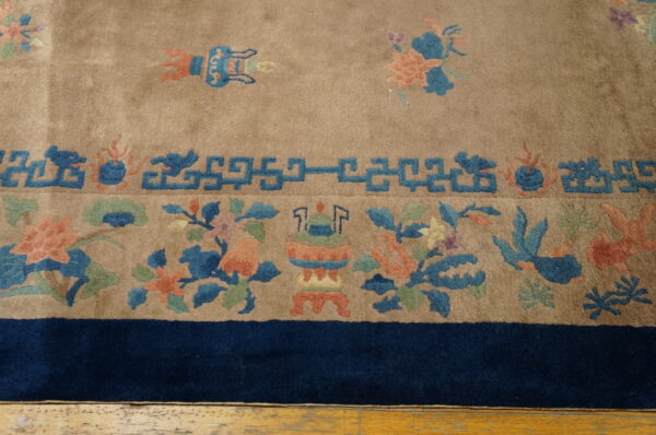 Tan rug border featuring blue geometric fretwork, coral floral motifs, and a solid navy edge on a wood floor.