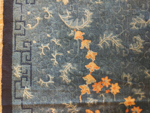 Textured blue rug displaying orange flowers, ivory vines, and a dark blue greek key border pattern.