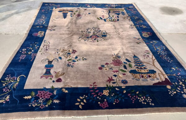 A large rug with a central beige field and scattered chinoiserie floral patterns, surrounded by a broad indigo border.