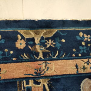 Detailed view of a navy blue rug border with tan, peach, and teal floral patterns on light tiled flooring.