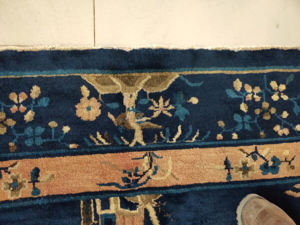 Detailed view of a navy blue rug border with tan, peach, and teal floral patterns on light tiled flooring.