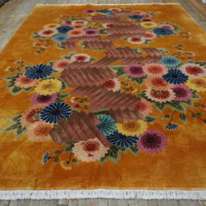 Velvety saffron rug with carved multicolored floral and brown geometric patterns rests on distressed wood flooring.