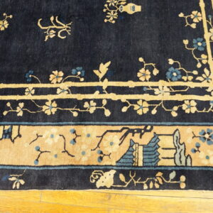 Detail of a dark blue rug featuring tan and teal floral patterns and architectural border on wood flooring.