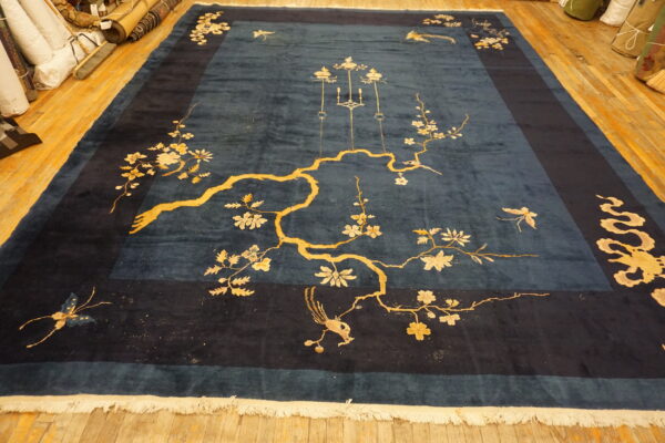 Large teal blue rug with a dark border, featuring a yellow flowering branch, birds, and butterflies on a wood floor.