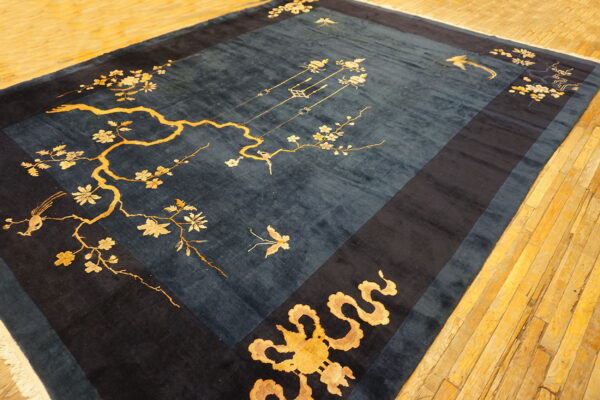 A dark blue rug with gold flowering branches and birds rests on a distressed yellow hardwood floor.