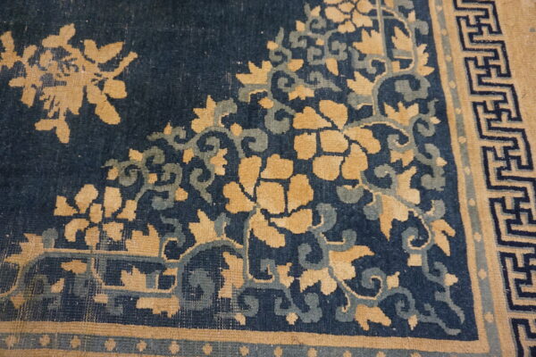 Dark blue rug corner showing golden tan flowers, teal vines, and a geometric fretwork border.