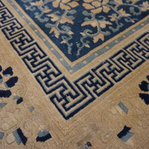 Tan and navy rug corner featuring a geometric fret border and floral motifs in the tan and blue fields.