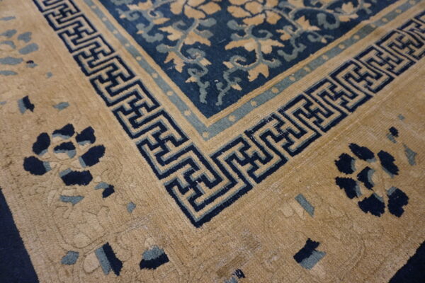 Tan and navy rug corner featuring a geometric fret border and floral motifs in the tan and blue fields.