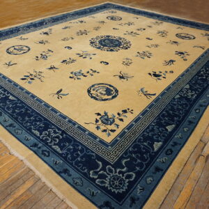 Square beige and blue patterned rug featuring a dense floral border sits on worn hardwood flooring.