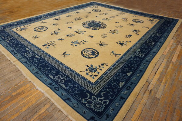 Square beige and blue patterned rug featuring a dense floral border sits on worn hardwood flooring.