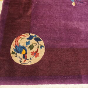 Magenta rug with a maroon border corner featuring a colorful bird medallion on distressed yellow wood.