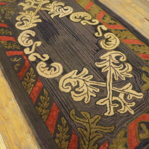 Hooked dark-toned rug featuring bold beige, red, and green stylized leaves on rustic wood plank flooring.