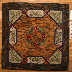 Textured hooked rug featuring a red floral wreath, clover corners, and an orange-brown center on wood planks.