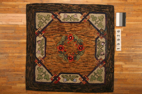 Textured hooked rug featuring a red floral wreath, clover corners, and an orange-brown center on wood planks.