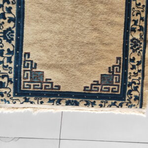 Close-up of a beige rug featuring deep blue borders and geometric fretwork in the corner on white tiles.