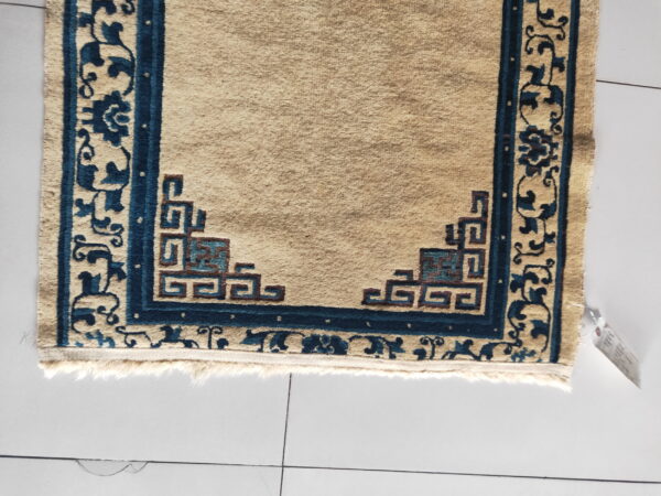Close-up of a beige rug featuring deep blue borders and geometric fretwork in the corner on white tiles.