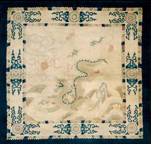 Pale square rug with faint blue and pink mythological figures and a prominent navy and cream scrolled border.