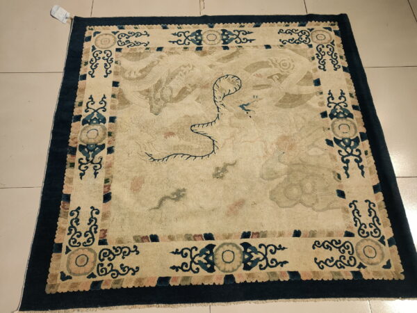 Cream rug with a central subtle dragon motif and a dark blue ornate border on light tile flooring.