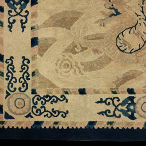 Tan rug corner showing low pile, scrolling borders, and stylized motifs in navy, teal, and pink.