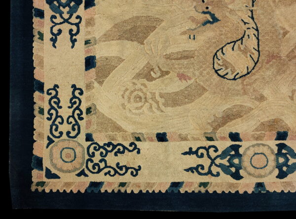 Tan rug corner showing low pile, scrolling borders, and stylized motifs in navy, teal, and pink.