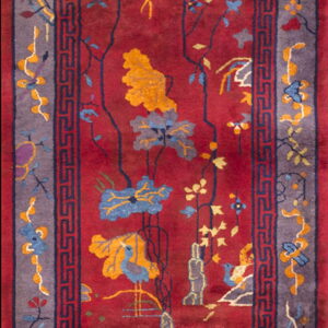 Rectangular rug with a red field featuring yellow and blue botanical scenes and bordered by a purple geometric trim.