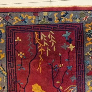 High-pile red rug with gray borders, dark blue key patterns, and colorful stylized floral motifs on white tile.