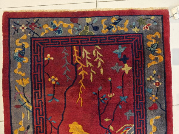 High-pile red rug with gray borders, dark blue key patterns, and colorful stylized floral motifs on white tile.