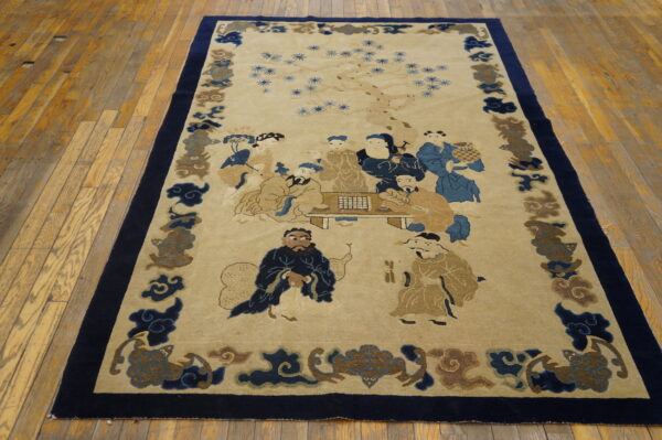 Low-pile beige rug with blue figures and a dark border, placed on worn yellow hardwood planks.