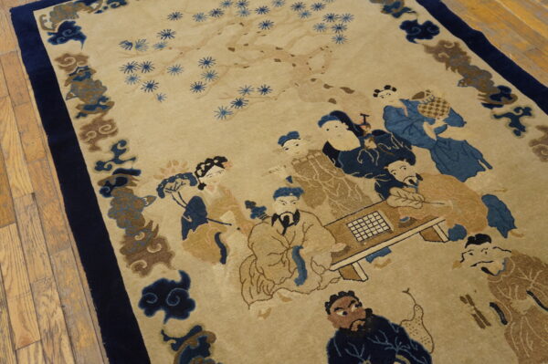 Low-pile beige rug with a blue and brown figural scene and wide navy border rests on wood flooring.