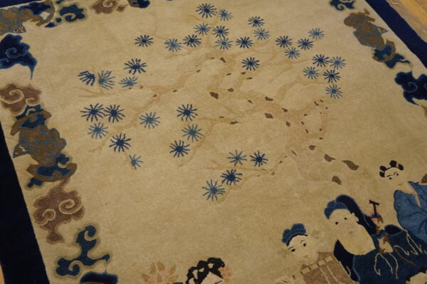 Beige field rug with blue floral sprays, stylized figures, and a dark blue and brown pictorial border.