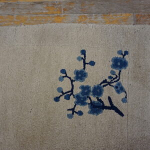 Textured beige rug with a blue blossom motif and fringe sits partially on distressed wood flooring.