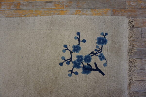 Textured beige rug with a blue blossom motif and fringe sits partially on distressed wood flooring.