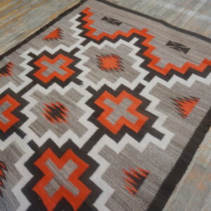 Geometric rug featuring stepped crosses and zigzags in orange, gray, black, and cream on distressed wood planks.