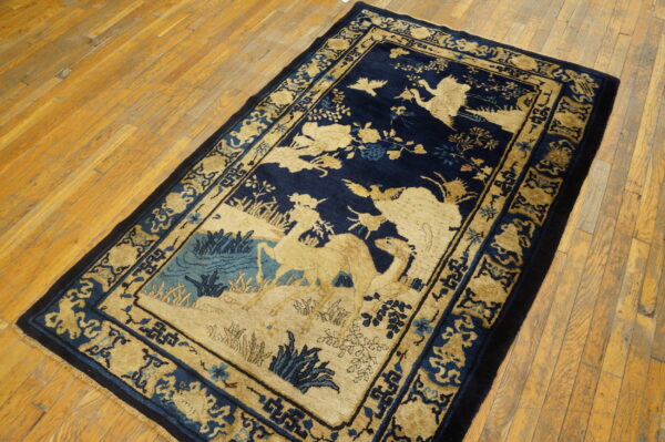 Navy and tan rug with a scenic pattern of animals and foliage, bordered by geometric motifs, on wood plank flooring.