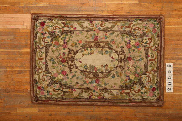 Beige hooked rug with dense, colorful floral patterns and brown baroque scrollwork on light hardwood.