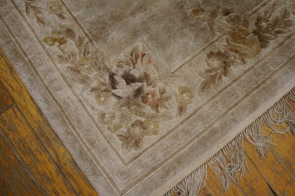 Muted beige floral rug featuring detailed carved flowers and a geometric border on wood flooring.