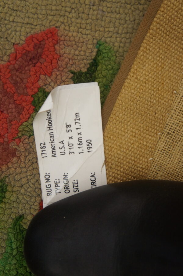 Hooked pile rug in olive, red, and green floral pattern, showing its woven backing and product label.