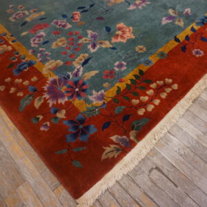 Teal field floral rug with a thick red border and fringe on distressed plank flooring.