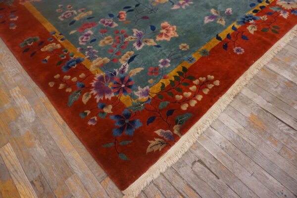 Teal field floral rug with a thick red border and fringe on distressed plank flooring.