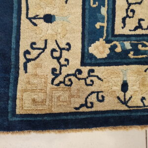 Textured tan and deep blue rug corner showing floral motifs and a key fret pattern rests on white tile.