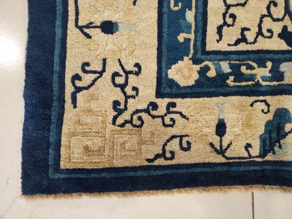 Textured tan and deep blue rug corner showing floral motifs and a key fret pattern rests on white tile.