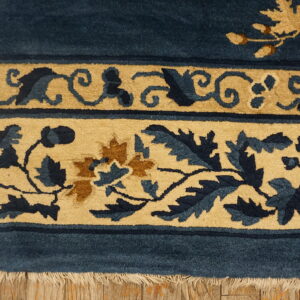 Deep blue rug with tan floral borders and visible fringe lying on distressed wood floor.