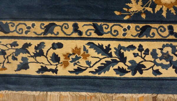 Deep blue rug with tan floral borders and visible fringe lying on distressed wood floor.