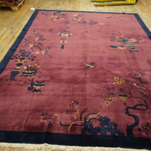 Large red rug with dark blue borders and traditional botanical patterns lies on light wooden floorboards.