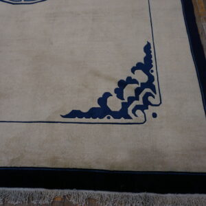 A light tan rug with dark blue stylized corner details and a wide border on distressed wood flooring.