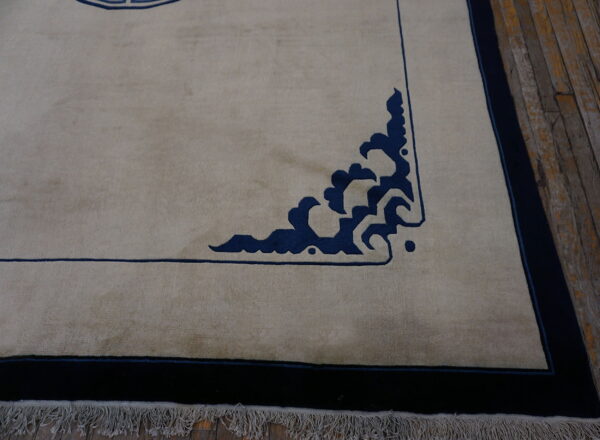 A light tan rug with dark blue stylized corner details and a wide border on distressed wood flooring.